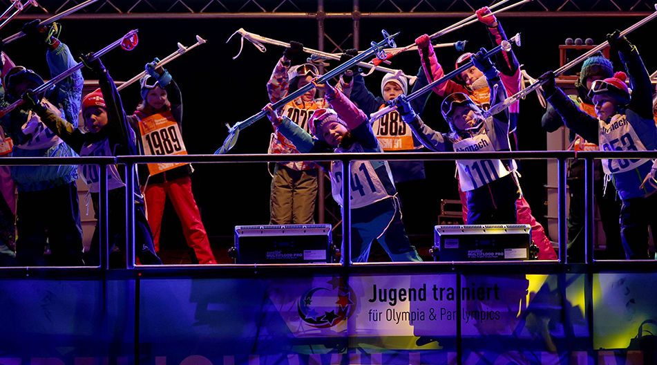 Eine Gruppe von Kindern in Wintersportkleidung steht auf einer Bühne und hält Skistöcke hoch, als würden sie gemeinsam trainieren oder jubeln.
