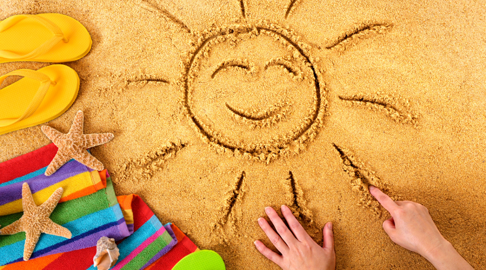 Lächelnde Sonne in den Sand gezeichnet, daneben Flip-Flops, Strandtuch und Muscheln.