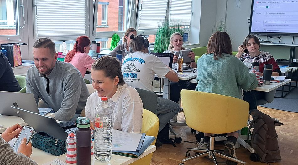Mehrere junge Erwachsene arbeiten in Kleingruppen an Tischen mit Laptops und Tablets in einem modernen Seminarraum des KBS. Auf einem Bildschirm im Hintergrund ist eine interaktive Lernplattform zu sehen. Die Stimmung wirkt konzentriert und kooperativ.