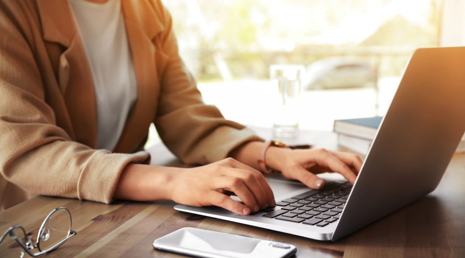 Person tippt auf Laptop-Tastatur an einem Holztisch mit Smartphone, Brille und Glas Wasser