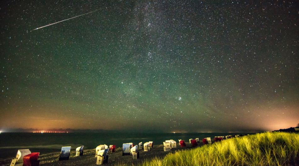 Ein Sternenhimmel über dem Meer. Am Strand stehen zahlreiche Strandkörbe, die durch den grünlich schimmernden Nachthimmel erleuchtet werden. Ein Sternenhimmel über dem Meer. Am Strand stehen zahlreiche Strandkörbe, die durch den grünlich schimmernden Nachthimmel erleuchtet werden.